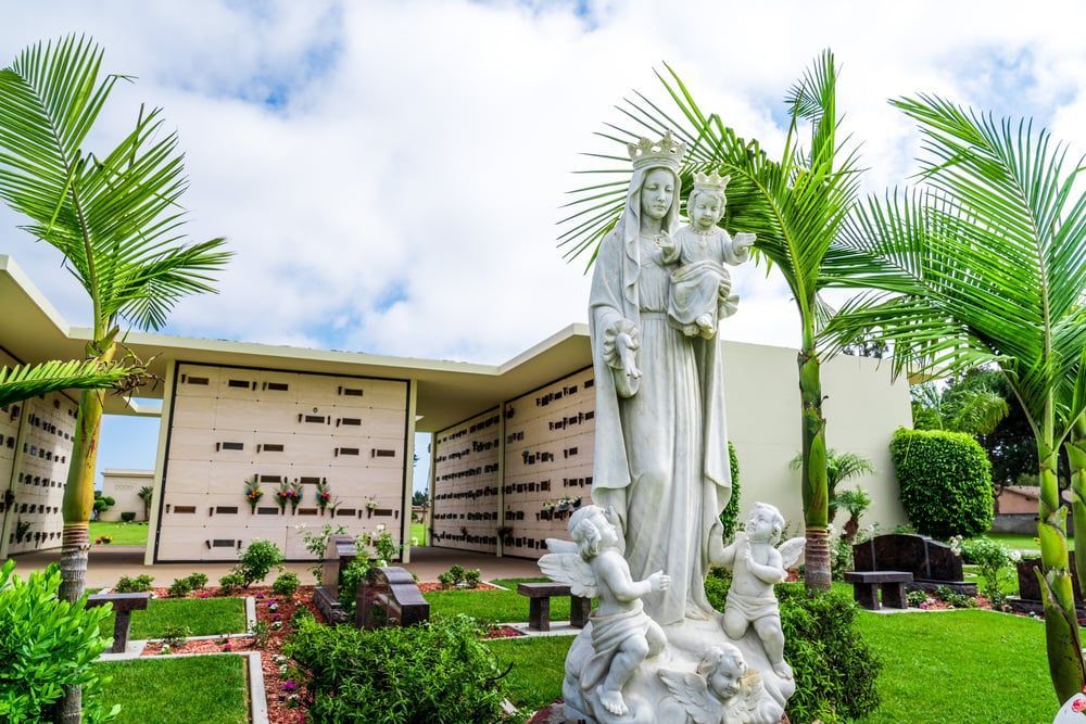 White statue among green plants in a landscaped memorial garden with plaques and walls in the background