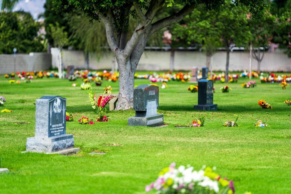 Grassy cemetery with gray headstones, colorful flowers, and large trees in the background