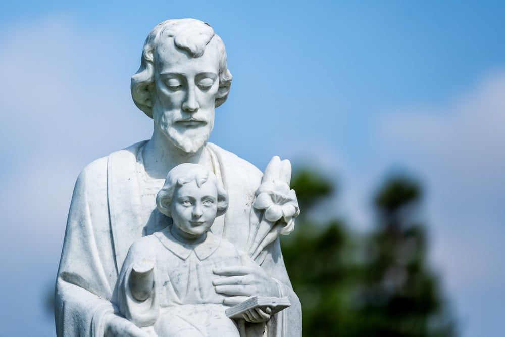 White statue of a man with a child in front, set against a blue sky and blurred trees