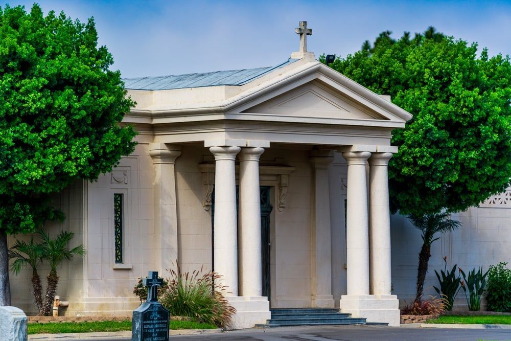 Small white mausoleum with columns and cross, framed by green trees in a cemetery