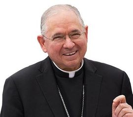 Smiling man in black clerical attire with a white collar, gesturing with one hand against a white background