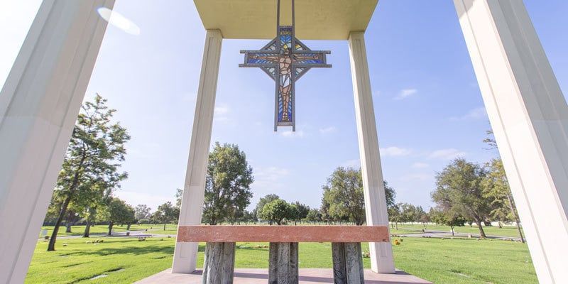 Crucifix hanging between white columns in a park, with a wooden bench and trees in the background