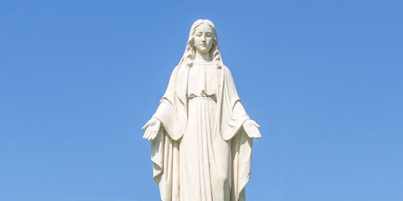 White statue of the Virgin Mary with outstretched arms against a blue sky
