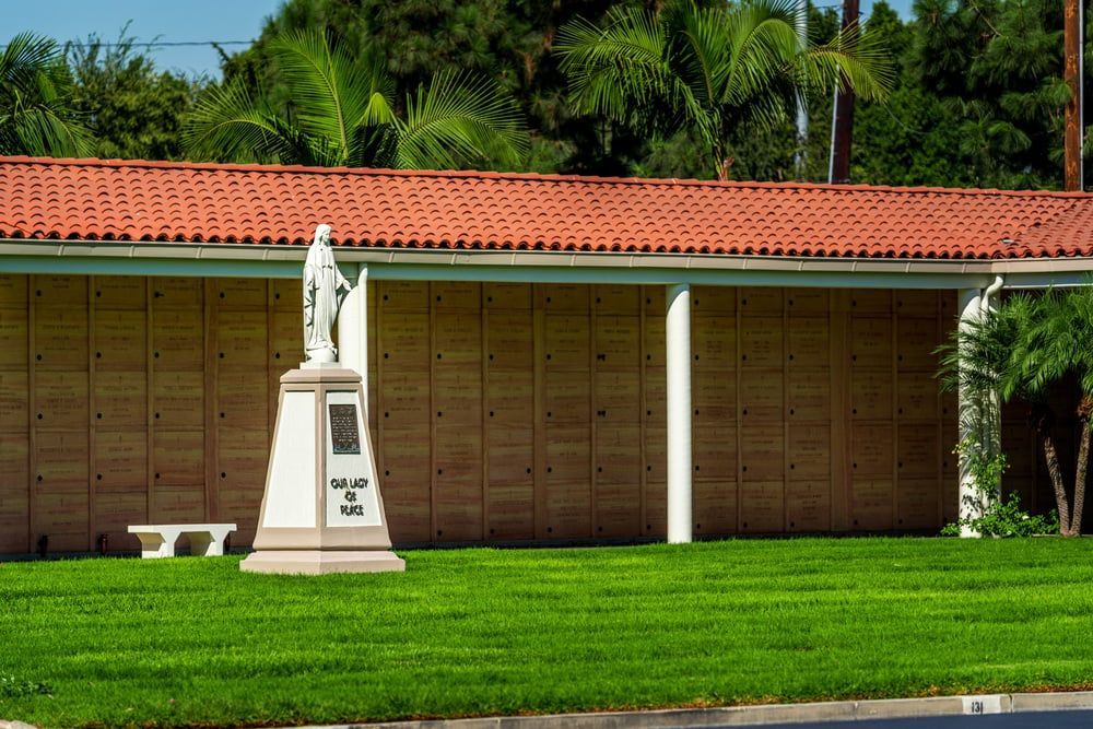 White statue on pedestal in front of a tan building with a red tile roof and green lawn.