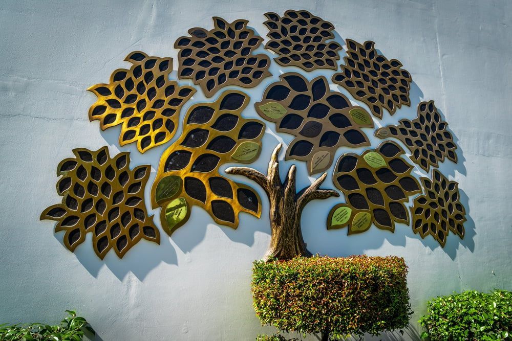 Decorative metal tree wall art with gold and black leaves above trimmed greenery on a gray wall