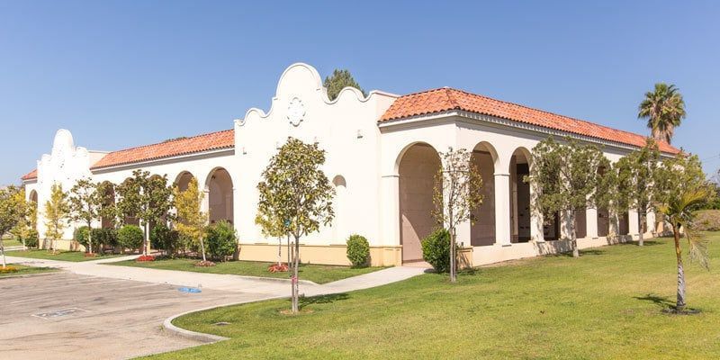 Spanish-style stucco building with red-tile roof, arched windows, and young trees on a sunny lawn