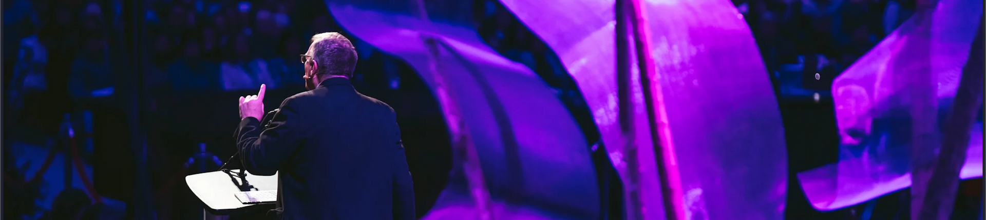 Person speaking on stage beside a large purple-lit curved structure, seen from behind.