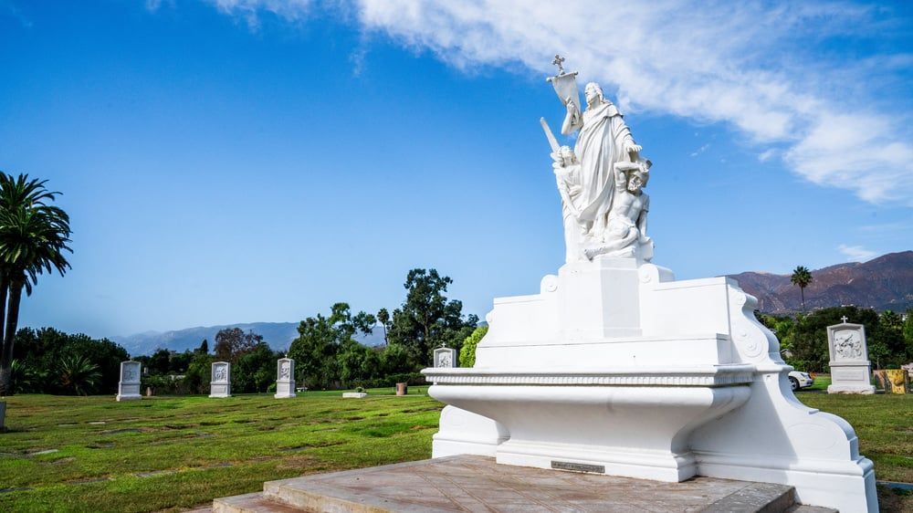 White statue on a pedestal in a grassy park with palm trees, mountains, and blue sky in the background