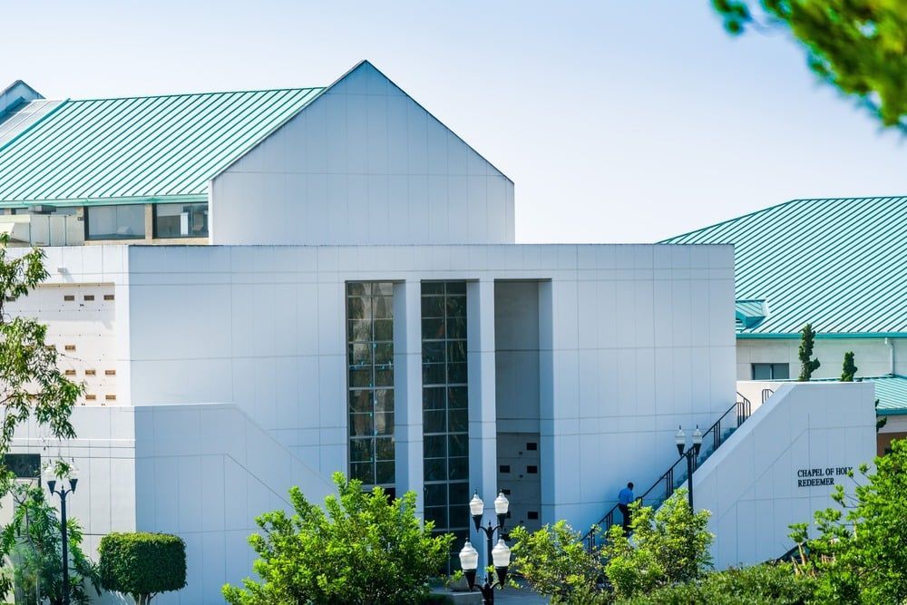 Modern white building with a teal roof and large windows, surrounded by trees and greenery