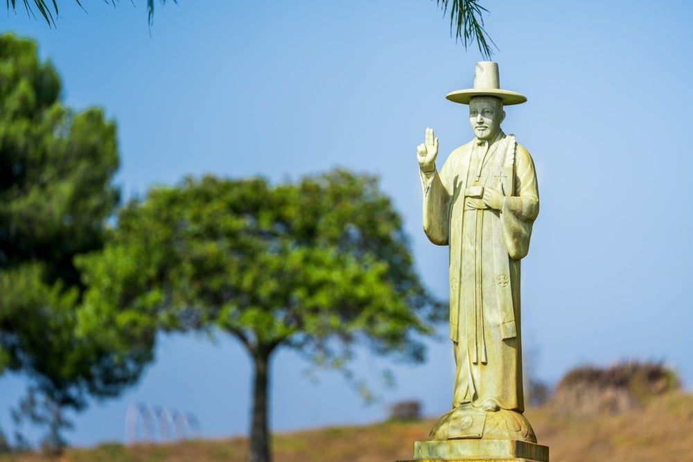 Statue of a robed figure in a hat, standing outdoors with trees and blue sky in the background