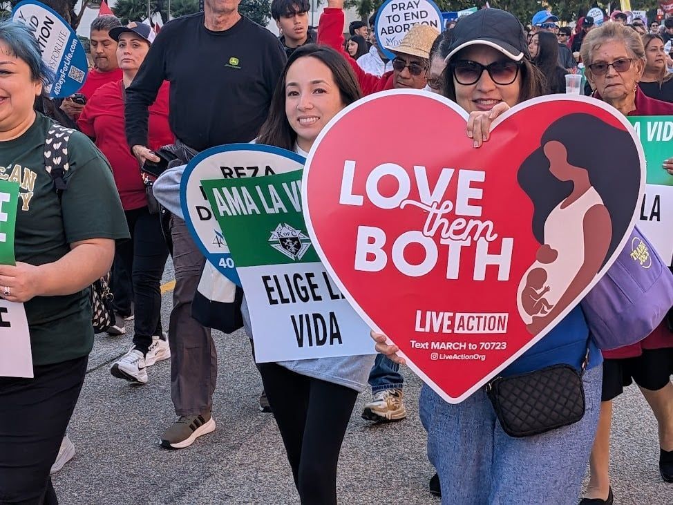 Protesters march holding signs, including a red heart reading “LOVE THEM BOTH” and “ELEGI VIDA.”