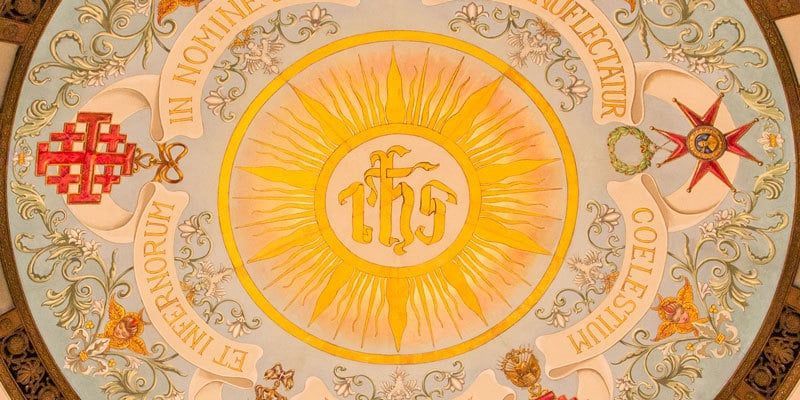 Golden religious emblem with sunburst, cross, and ornate circular Latin inscription on a patterned background