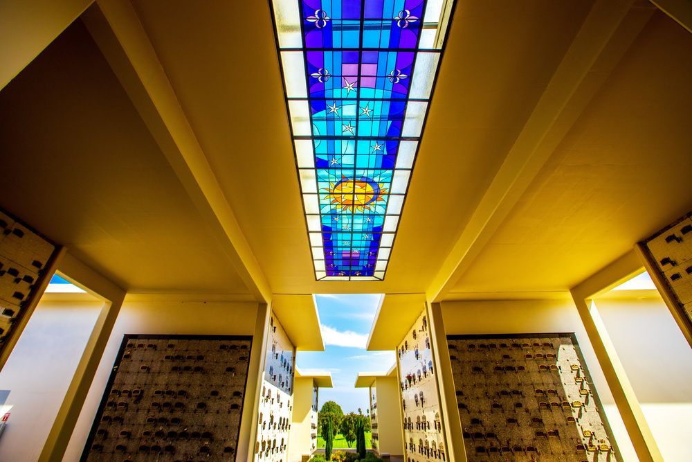 Stained-glass ceiling above a yellow-walled corridor with patterned pillars and a view of trees outside