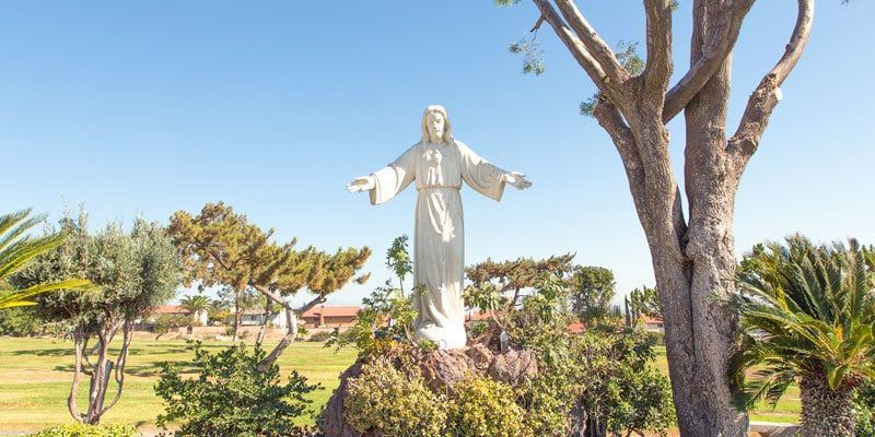 White statue with outstretched arms in a sunny garden beside trees and shrubs