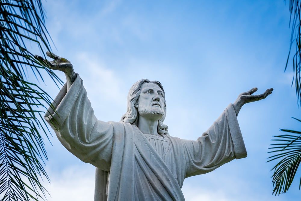 White statue of Jesus with raised arms against a blue sky, framed by palm leaves