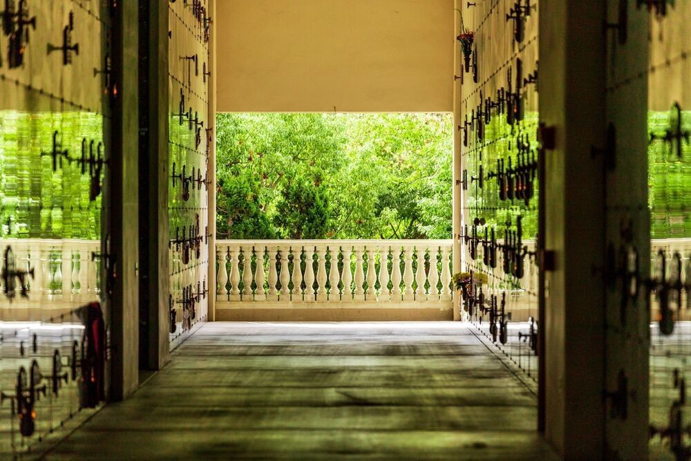 Hallway opening to a bright green garden, with ornate iron doors and a balcony railing.