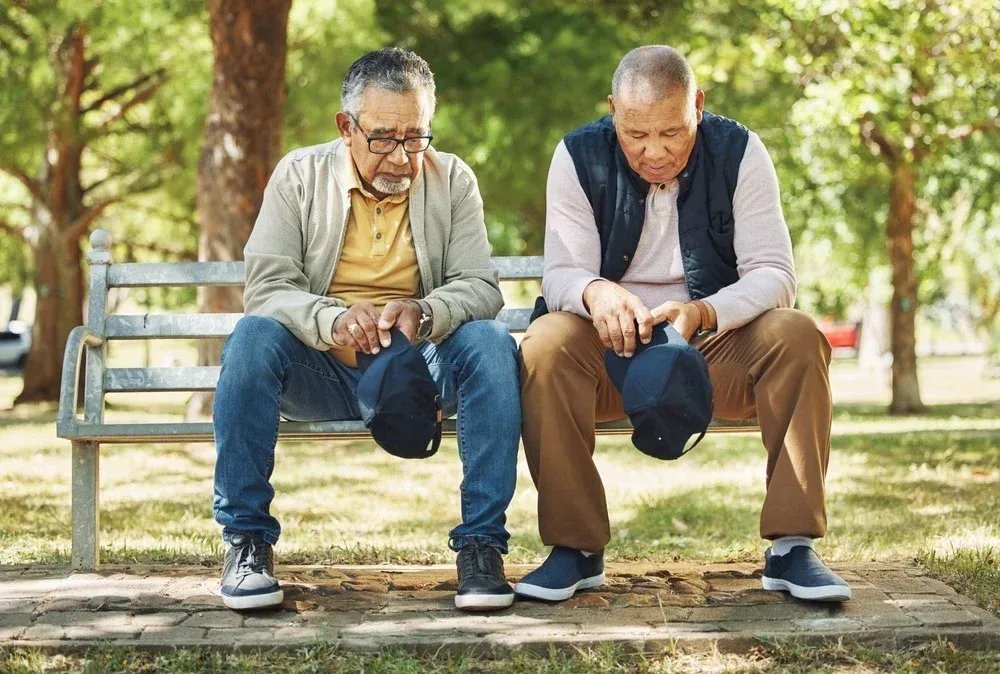 Two men sit on a park bench, looking down at their shoes in a sunny, tree-lined park.