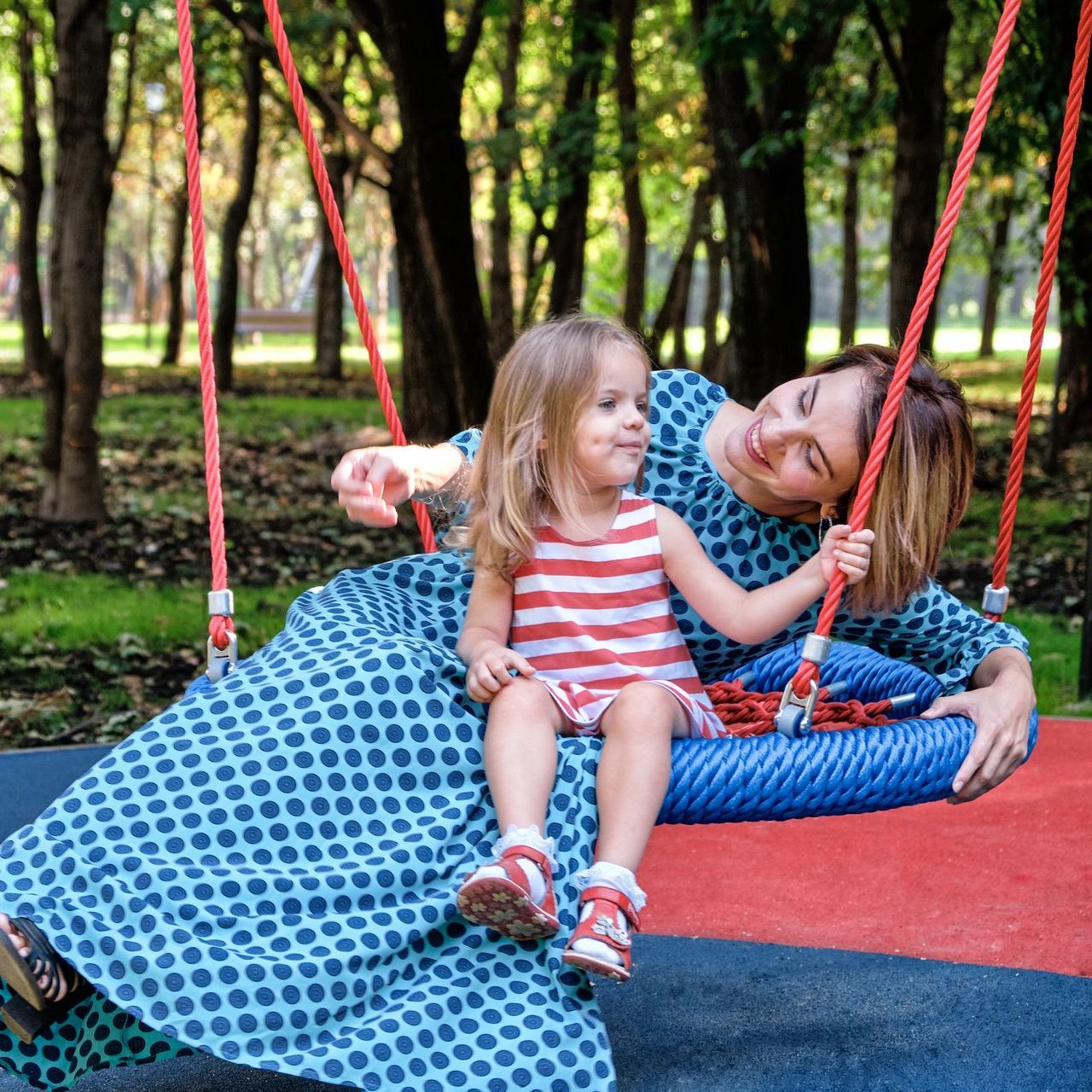 Woman and child on a swing in a park. Woman wearing blue dress, child wearing striped dress, both smiling.