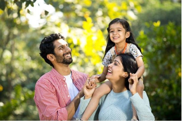 A smiling parent carries a child on their shoulders in a park, while another adult looks on with a joyful expression.