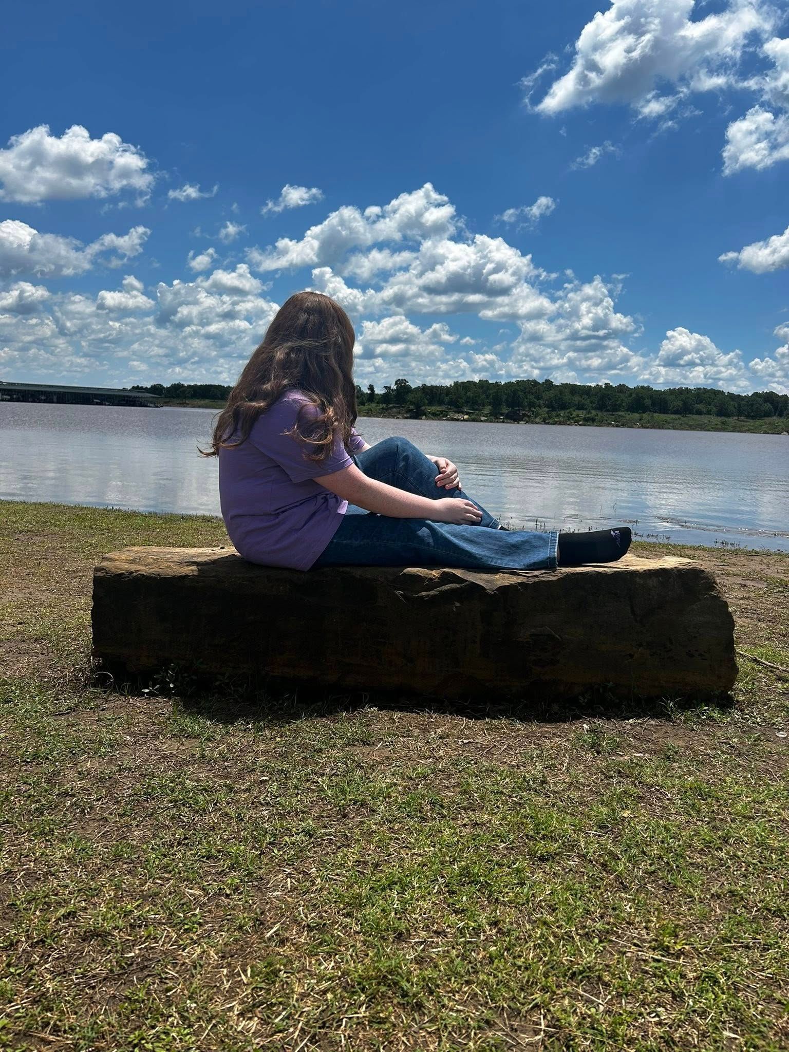 Woman sitting on a log, looking at a river under a blue sky with clouds.