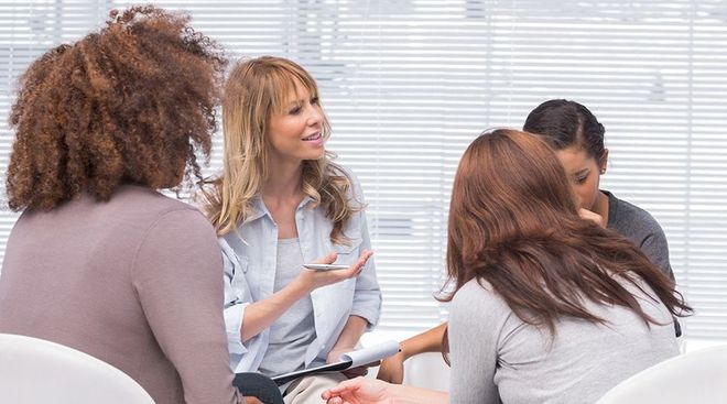 Four women in a circle, one speaking while others listen. Indoor setting with blinds.