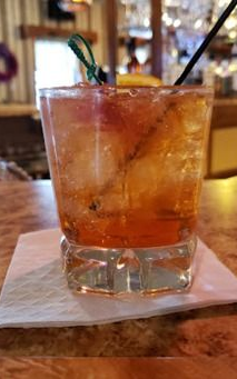 Cocktail in a clear glass on a bar, garnished with a green sprig and orange slice.