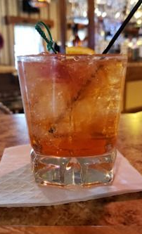 Cocktail in a clear glass on a bar, garnished with a green sprig and orange slice.