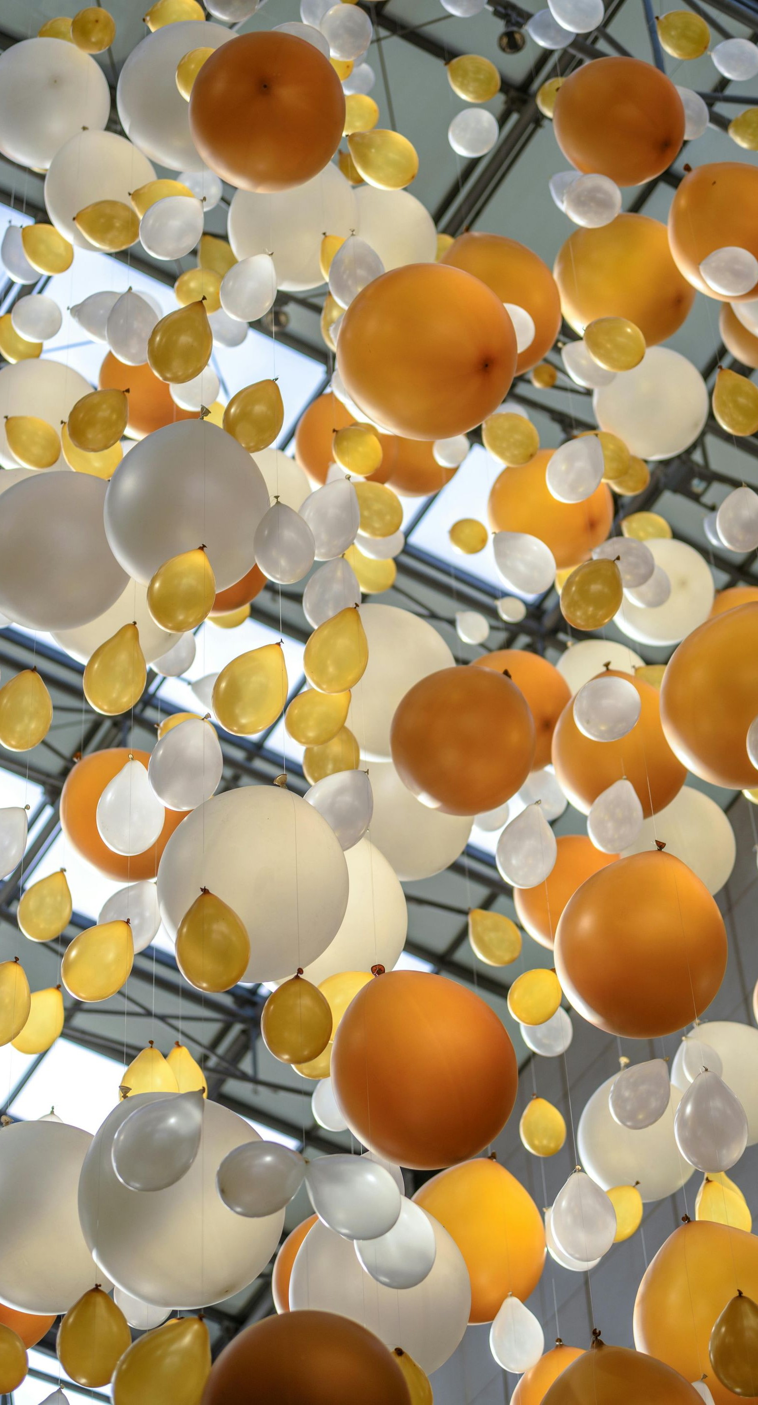 White and orange balloons floating in a glass-roofed area. Some balloons have gold leaf accents.
