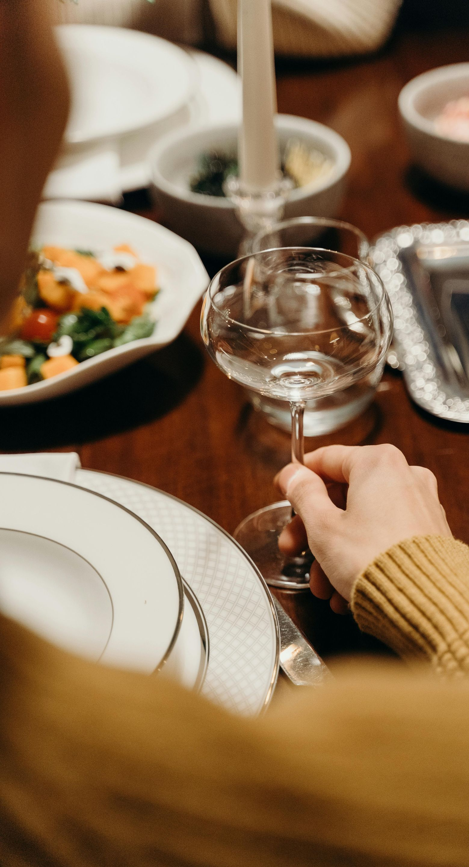 Person holding a wine glass at a dinner table set with plates, food, and candles.