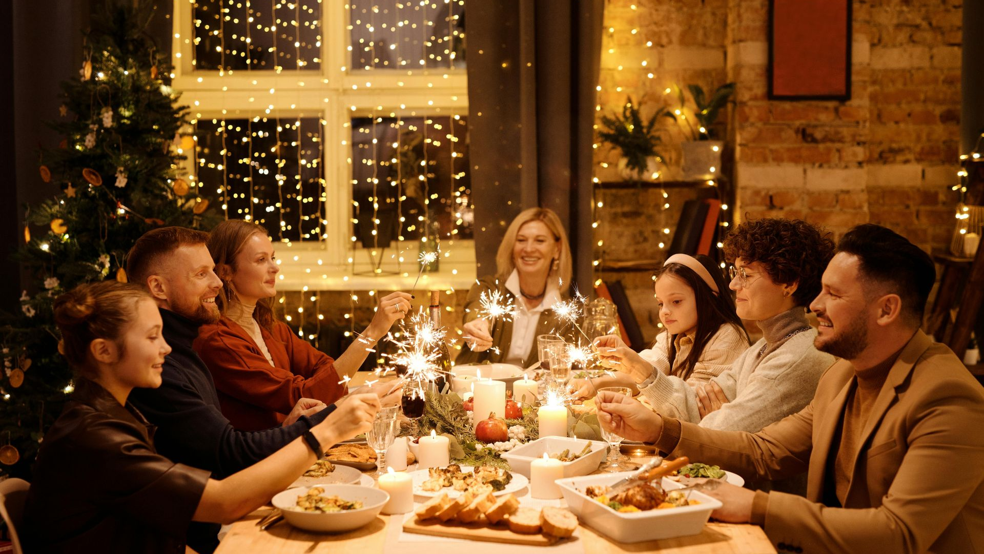 Family celebrating a holiday, seated around a table with food, holding sparklers, festive decor.