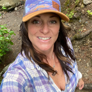 Woman smiles, wearing a plaid shirt and hat, hiking on a trail.