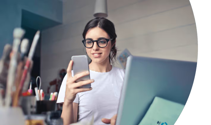 Woman with glasses smiling at phone beside a laptop in a bright office