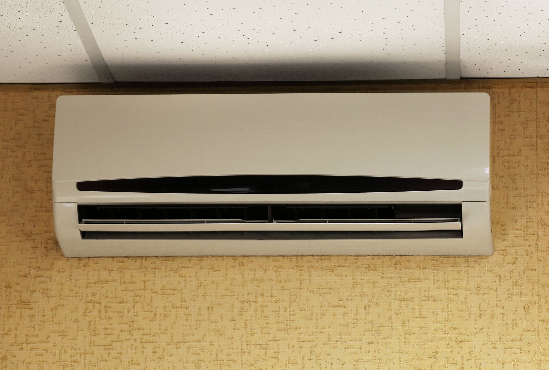 Wall-mounted white air conditioner on a beige wall beneath ceiling tiles.