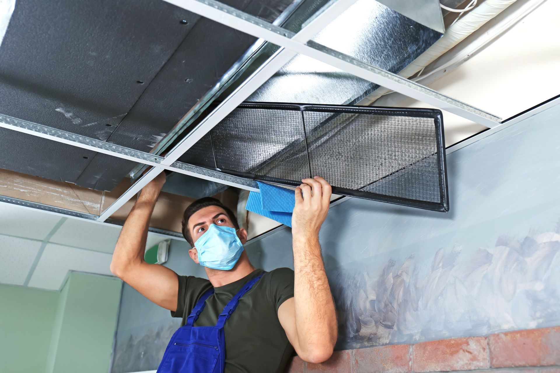 A man wearing a mask is cleaning a duct from the ceiling.
