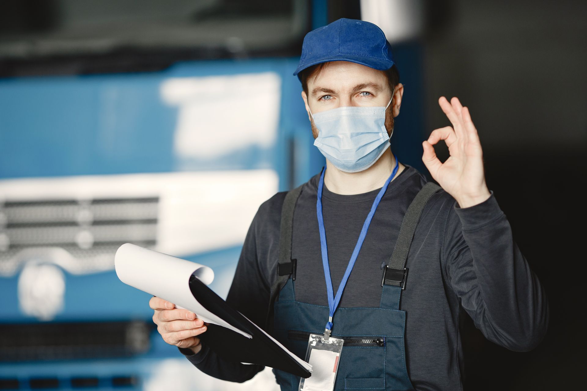 Mechanic in blue cap and mask holding a clipboard, making an OK gesture in front of a truck