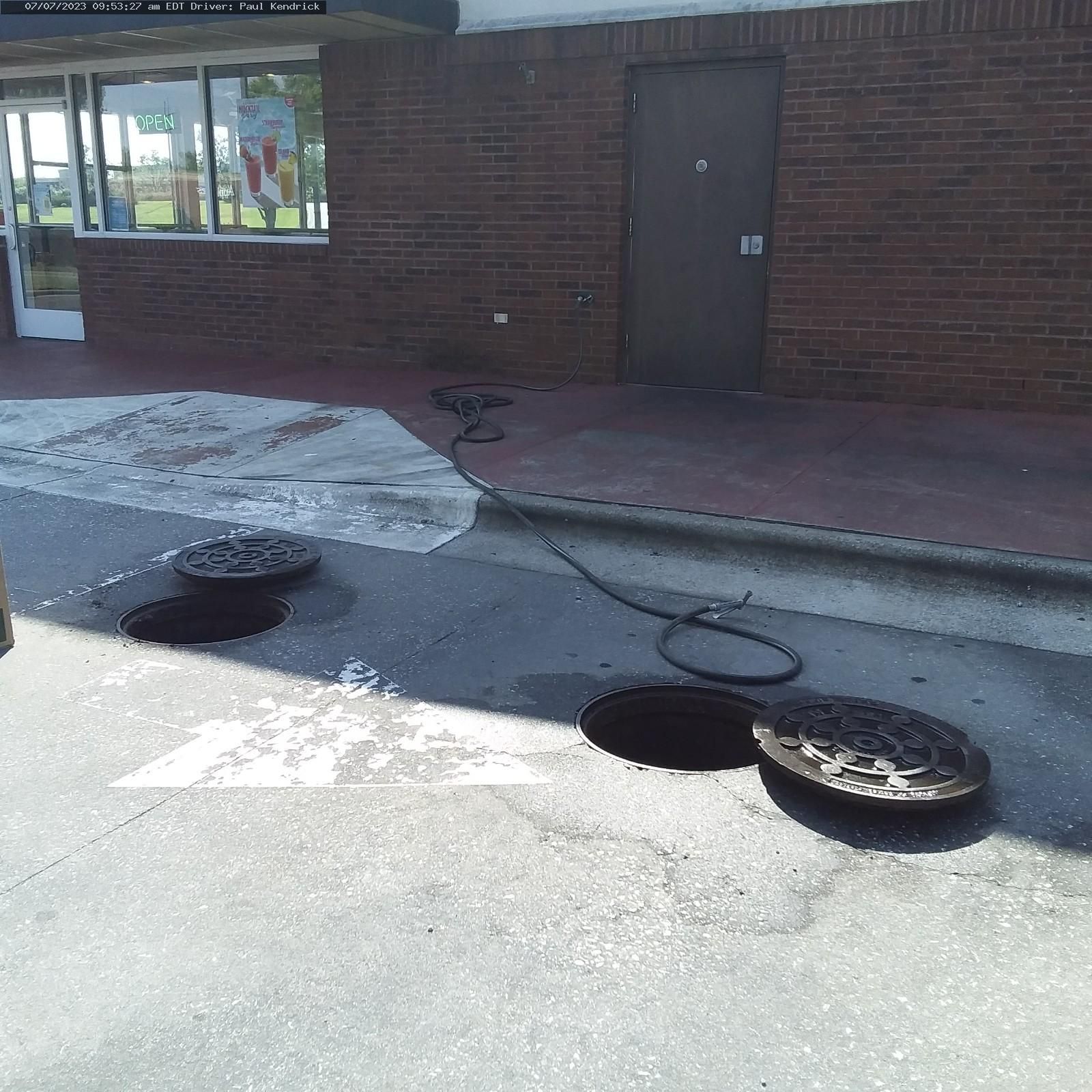 Two open manholes in an asphalt parking lot outside a brick building, with a black hose extending from one into a doorway.