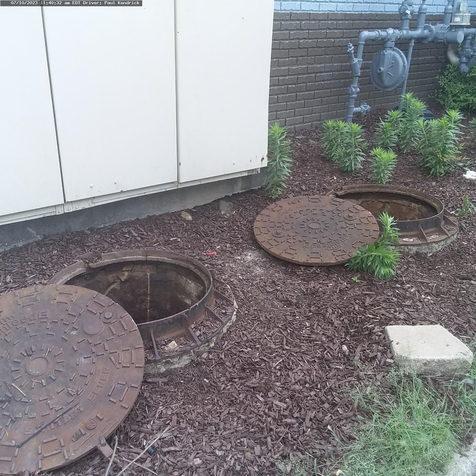 Two circular, rusted metal manhole covers lie open on a bed of dark mulch next to a building and gas utility equipment.
