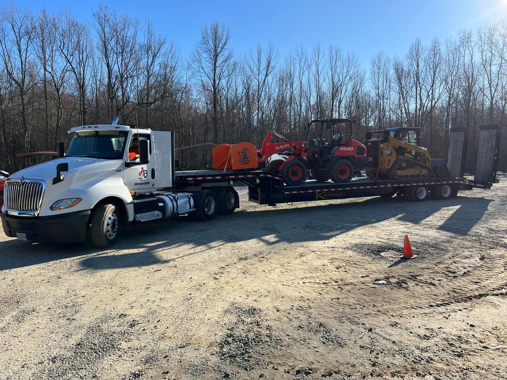A white semi-truck hauls a flatbed trailer loaded with a red compact tractor and a yellow skid-steer on a gravel lot.