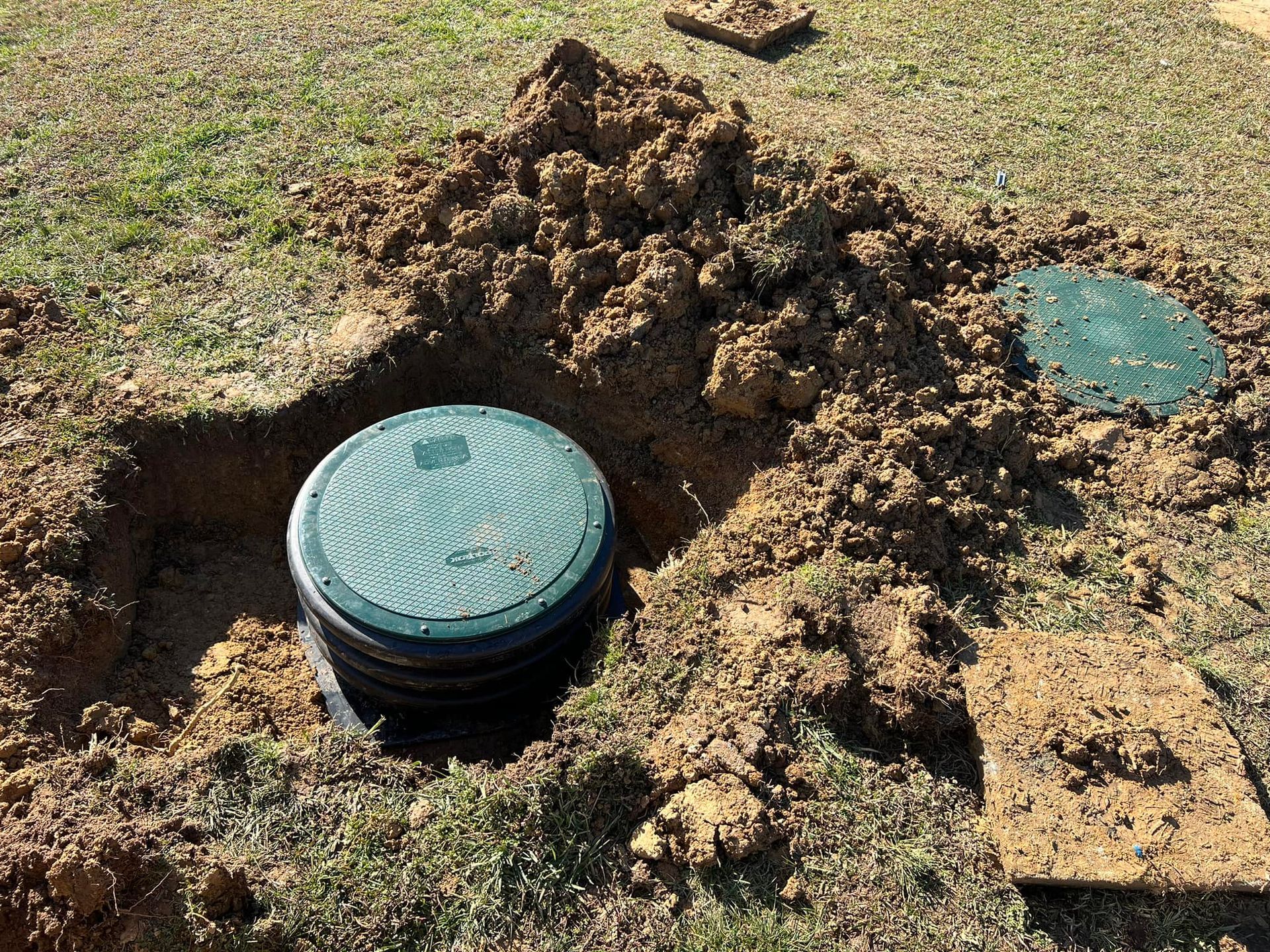 Two green circular septic tank access covers set in the ground, one partially excavated and surrounded by loose dirt.