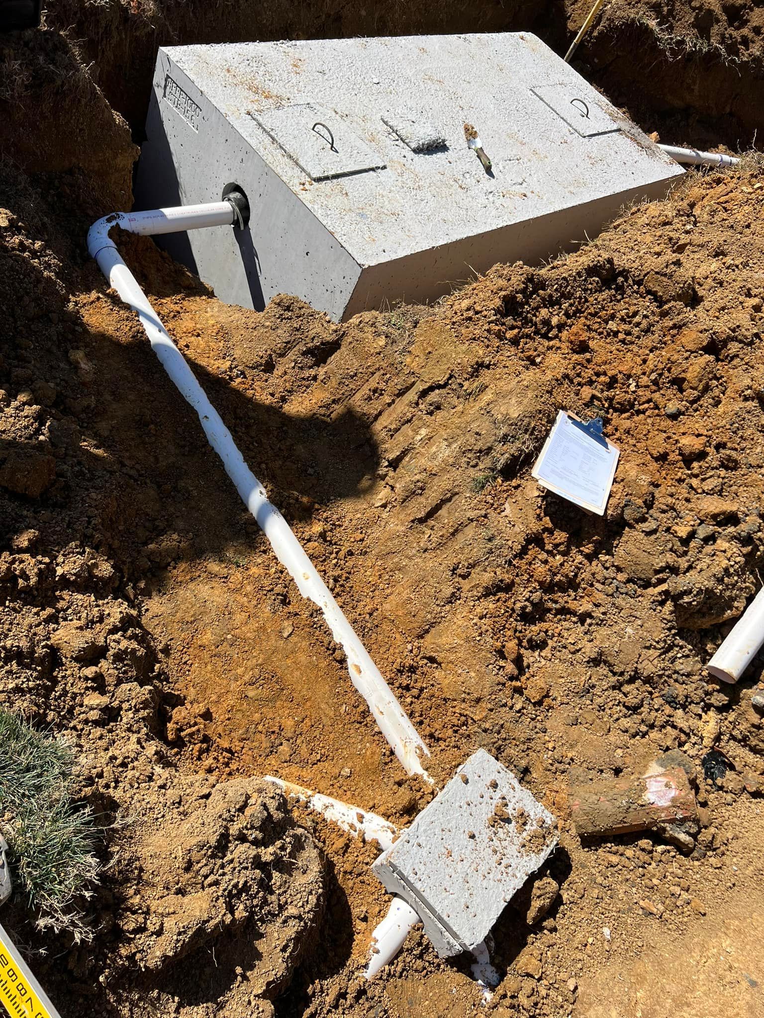 A partially buried concrete septic tank connected to white PVC pipes in a dirt construction site.