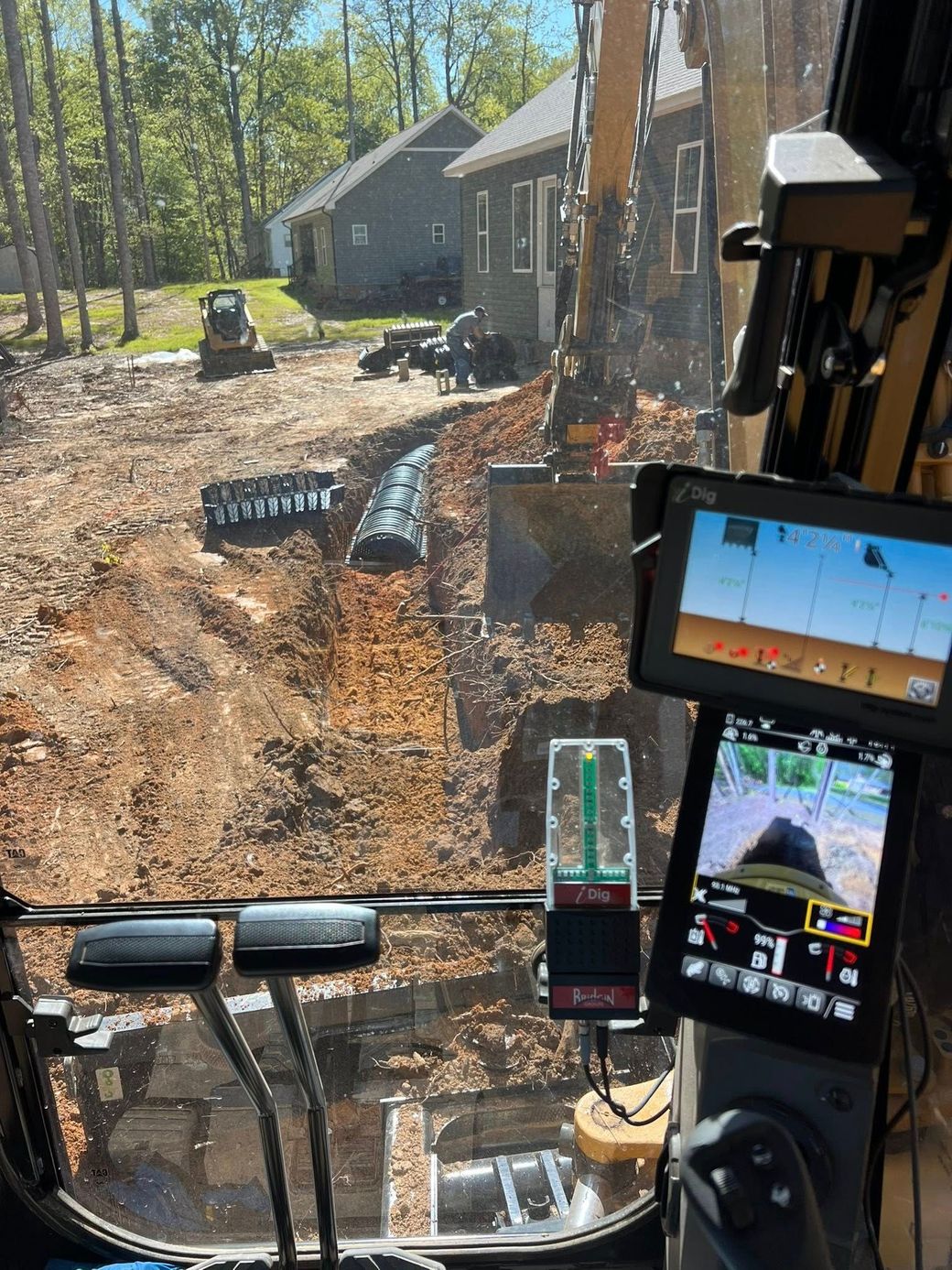 View from inside an excavator cab showing a construction site, a house in the distance, and machine monitoring screens.