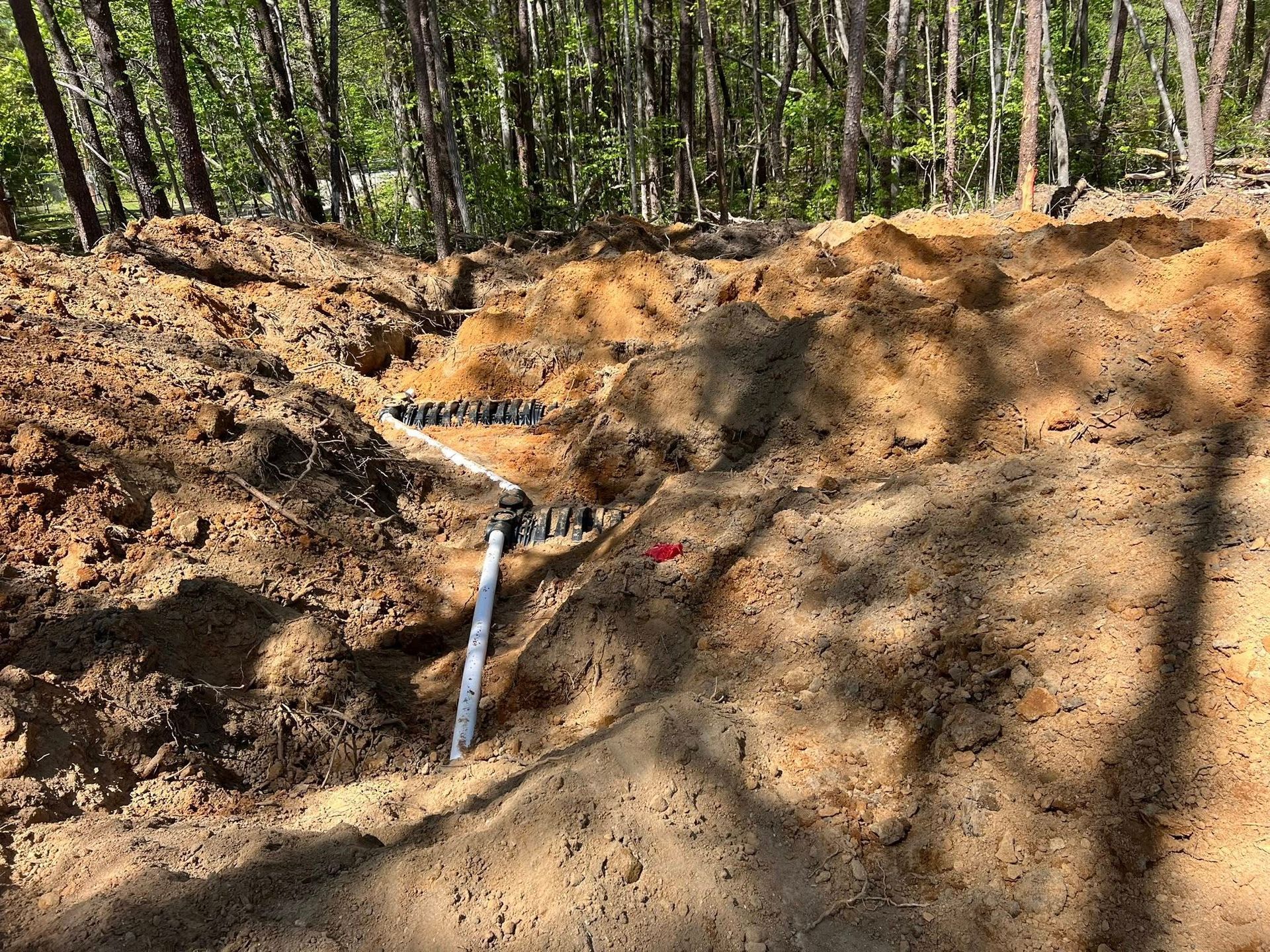 A white PVC pipe and dark drainage components installed in a trench dug into sandy soil, with woods in the background.