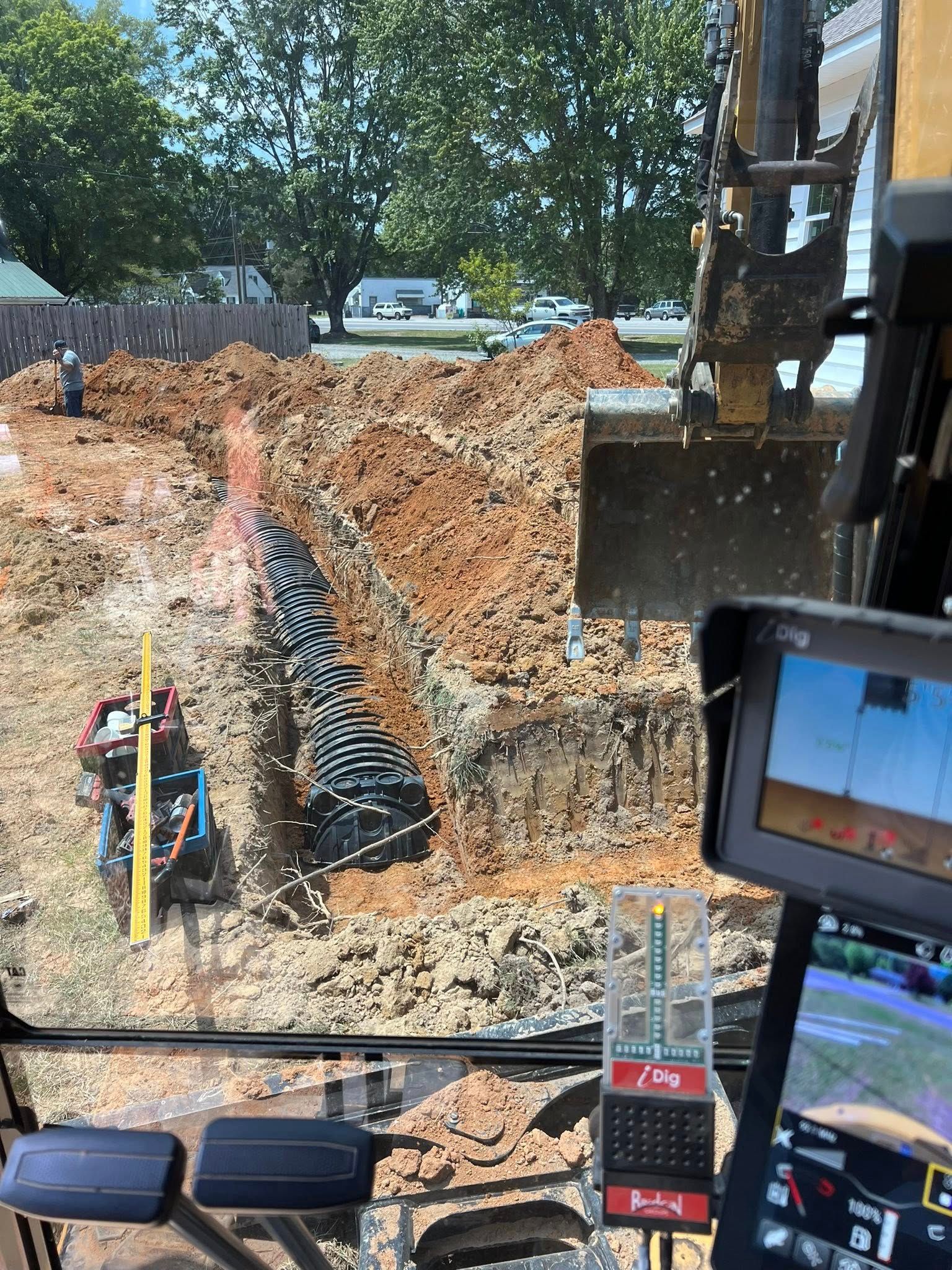View from inside an excavator cab showing a trench with a black drainage pipe and a laser level on a dirt construction site.