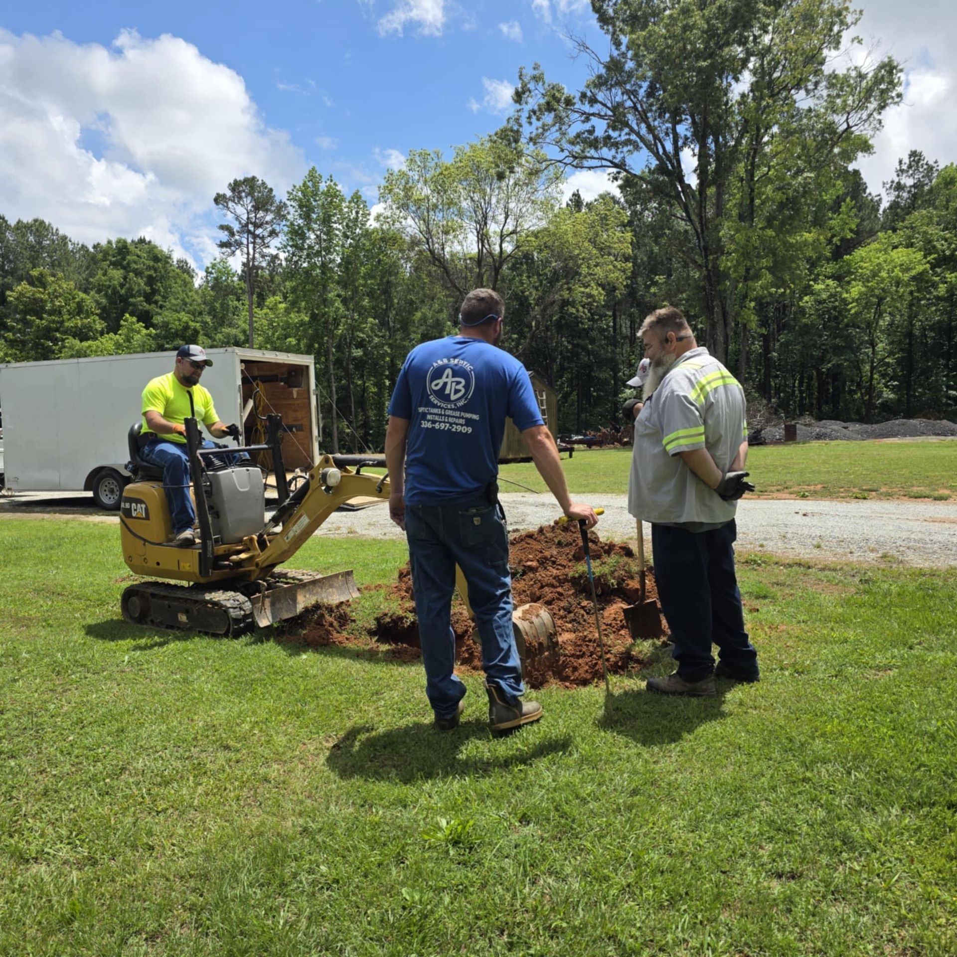 Two workers stand over a small excavation site in a grassy field, while a third sits on a yellow mini-excavator nearby.