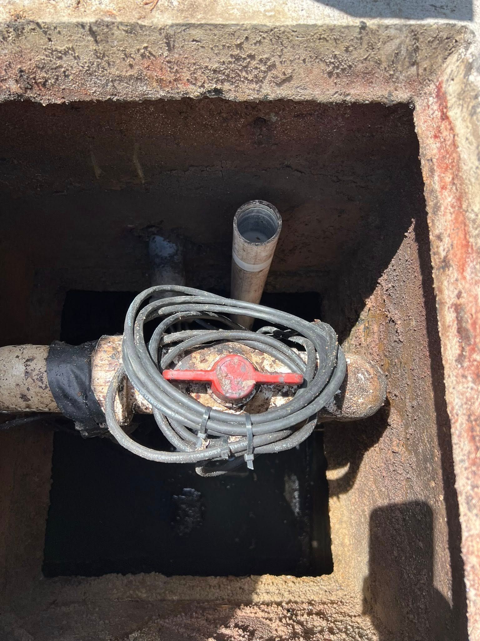 A metal utility pipe with a red shut-off valve and coiled black wire inside a recessed concrete box in the ground.