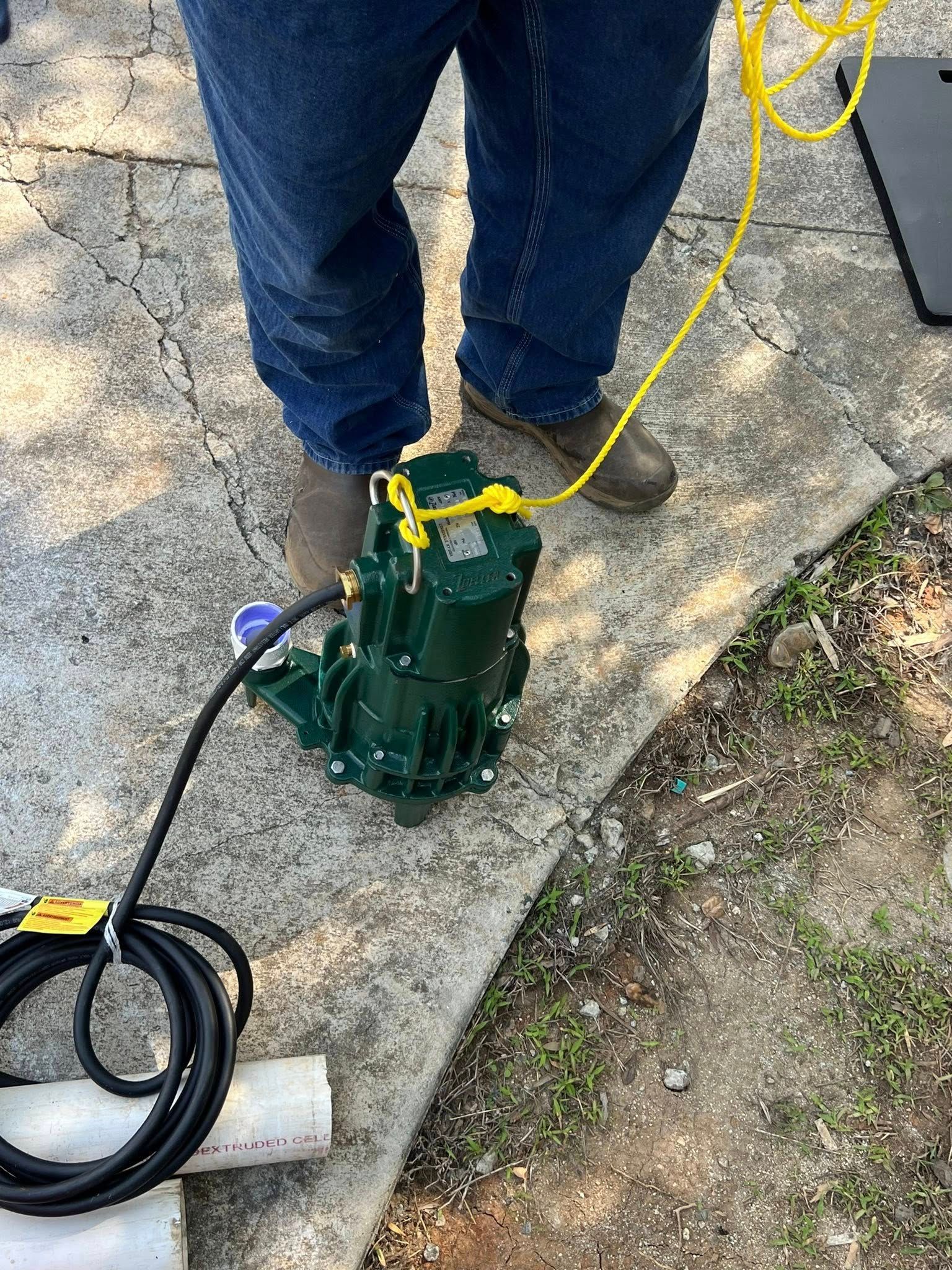 A person in blue jeans stands outdoors next to a dark green submersible pump attached to a yellow rope and black cord.