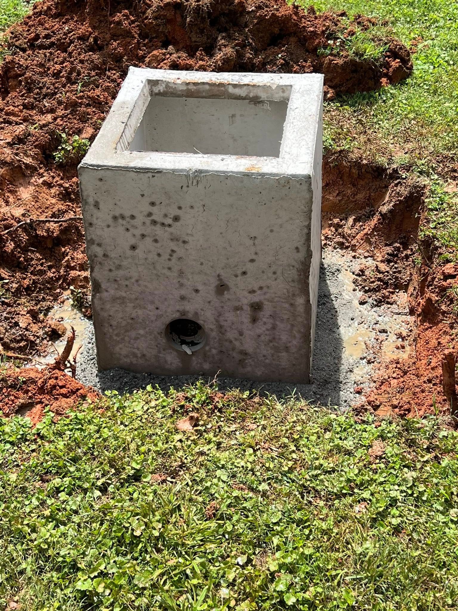 A square, grey concrete drainage catch basin sits in a hole in the ground surrounded by dirt and grass.
