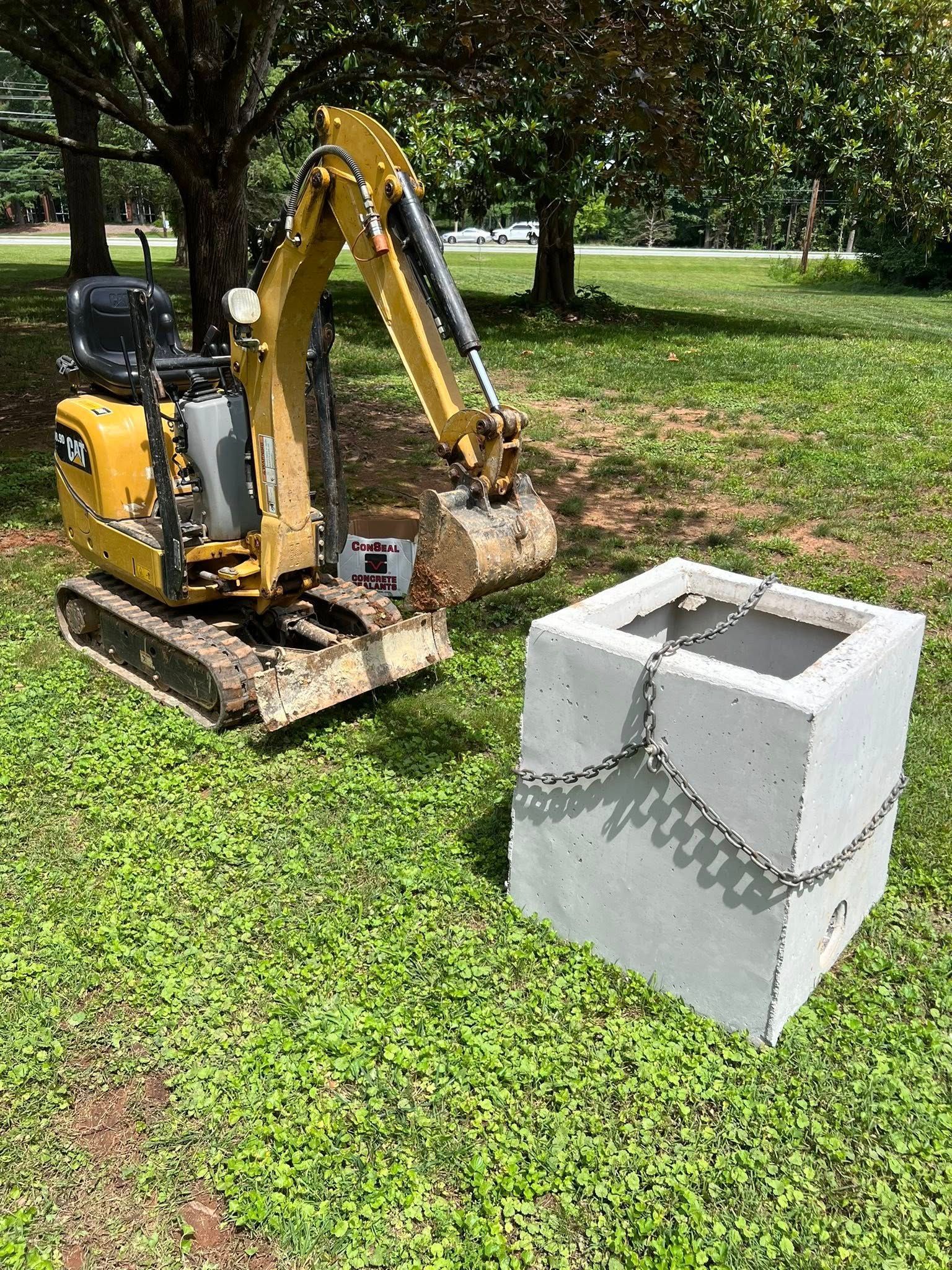 A yellow mini excavator sits on a grassy area next to a large, square concrete drainage structure wrapped in chains.