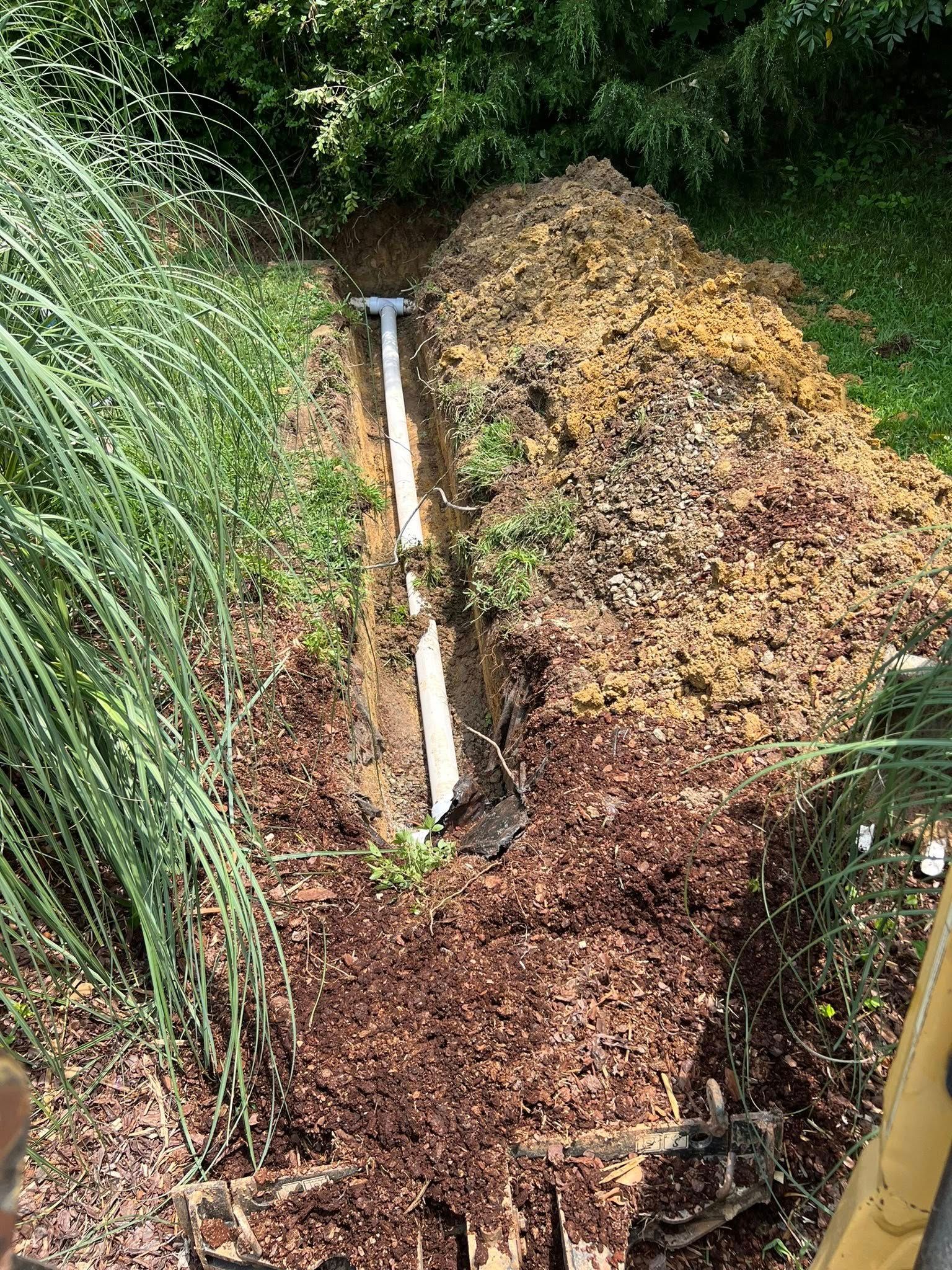 A white PVC pipe lies in a narrow, freshly dug trench alongside a pile of excavated dirt and mulch in a garden setting.