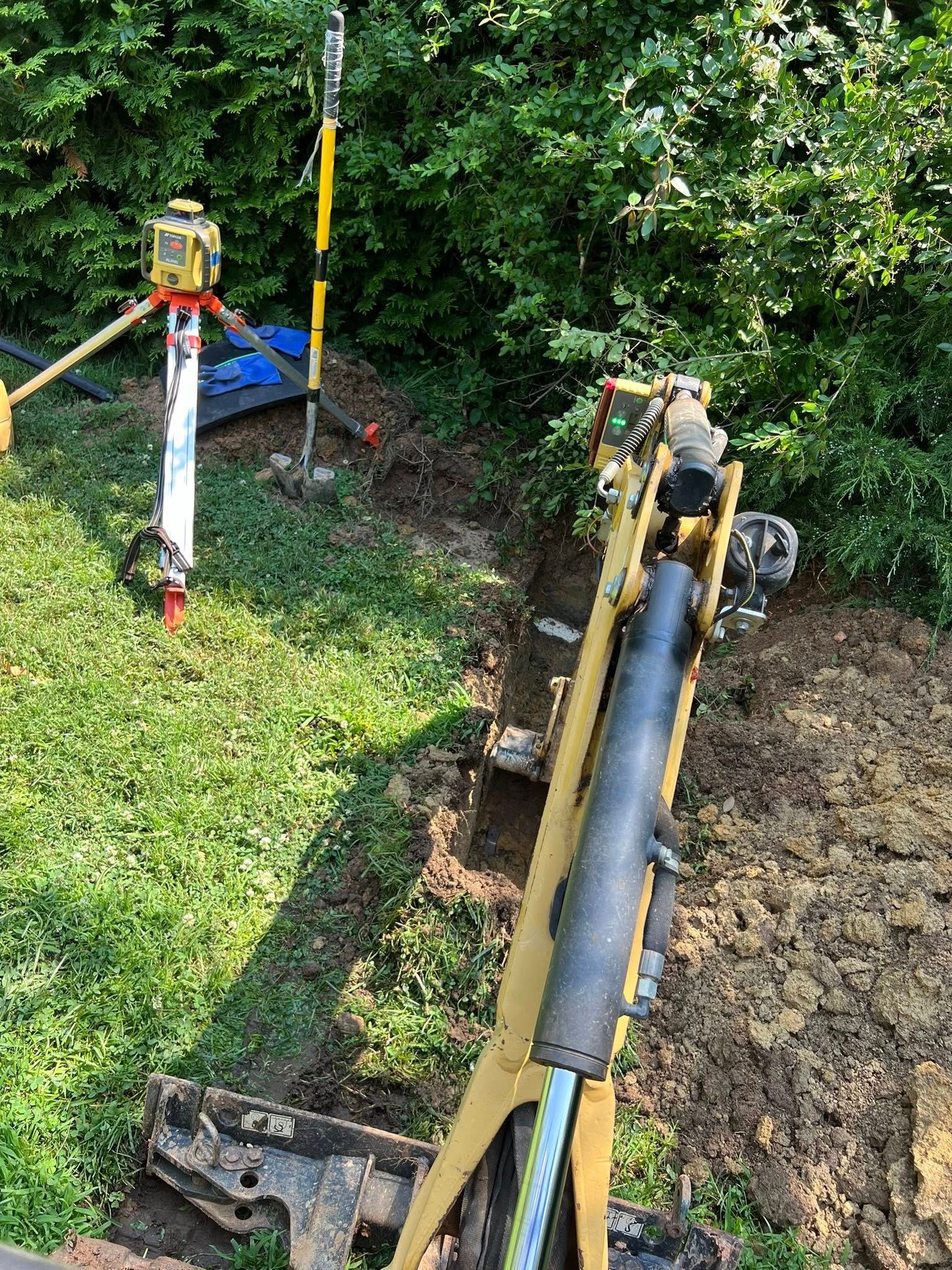 A small yellow excavator digging a trench near a surveying laser on a tripod in a grassy yard.